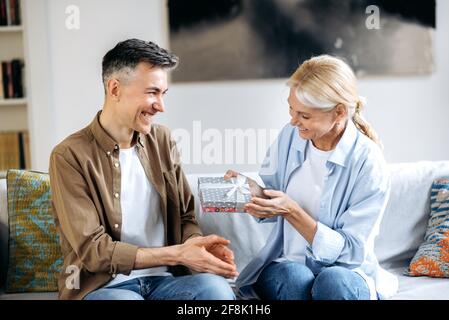 Ein Ehepaar mittleren Alters heiratete ein glückliches kaukasisches Paar zu Hause auf der Couch. Ein liebevoller Ehemann gibt seiner schönen Frau ein Geschenk, kleine Geschenkbox zu Ehren eines Geburtstags, Jubiläums oder März 8, sie glücklich zusammen Stockfoto