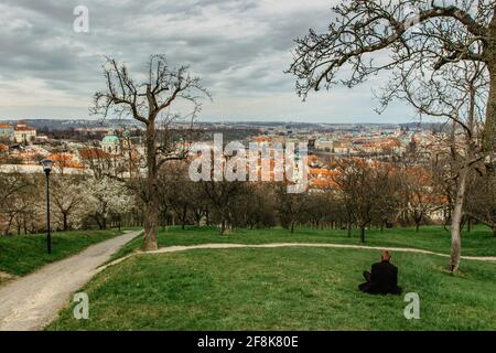 Einsamer Mann, der sich im Park entspannt und den Panoramablick auf Prag, Tschechien, genießt. Blühende Sakura-Kirschbäume auf dem Petrin-Hügel. Rote Dächer Stockfoto