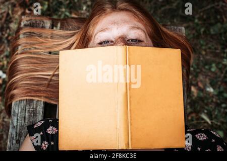 Draufsicht auf ein Teenager-Mädchen, das auf der Bank liegt und ein Buch über ihr Gesicht hält Stockfoto