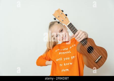 Das Teenager-blonde Mädchen in blauem T-Shirt hält ein hellbraunes Ukuleleinstrument Stockfoto