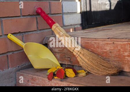 Ein Stapel herbstlicher roter und gelber Blätter, ein Besen und eine gelbe Kehrschaufel, die auf einer hölzernen Veranda gegen eine Ziegelwand mit abblätternder brauner Farbe am stehen Stockfoto