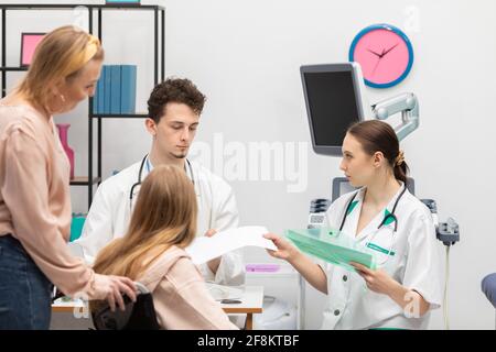 Ein im Rollstuhl sitzender Teenager und ihre Mutter warten in einer Arztpraxis. Die Krankenschwester gibt dem Arzt die Testergebnisse des Teenagers Stockfoto