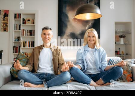 Gesunder Lebensstil. Verheiratetes glückliches Paar mittleren Alters verliebt, Ehemann und Ehefrau in legerer Kleidung, Yoga zu Hause auf dem Sofa, sitzend in Lotusposition im Wohnzimmer, meditierend, schauend auf die Kamera lächelnd Stockfoto