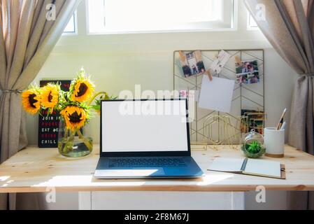 Arbeitsplatz im Home Office. Holzschreibtisch mit leerem weißen Laptop-Bildschirm, Moodboard mit pinierten Notizen und Fotos, Blumenstrauß in der Vase. Sonnig Stockfoto