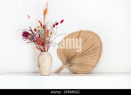 Buntes getrocknetes Blumenstrauß in einer hölzernen Vase im Boho-Stil, die auf einem weißen Regal mit einem Fächer aus getrocknetem Pam-Blatt daneben steht. Stockfoto