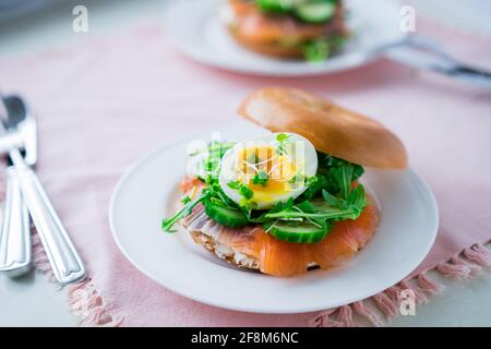 Frische Bagel-Sandwiches mit Lachs, weichem Ei, Salat, mikrogrünen Sprossen, Gurke und Frischkäse auf rosa Serviette mit Besteck auf weiß serviert Stockfoto