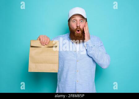 Deliveryman mit wunderten Ausdruck bereit, Beutel mit Lebensmitteln zu liefern. Cyan Hintergrund. Stockfoto