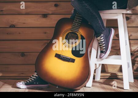 Akustische sechssaitige gelb-braune Gitarre und Beine einer Frau, die auf einem Hocker in Jeans sitzt, in der Nähe einer Holzwand Stockfoto