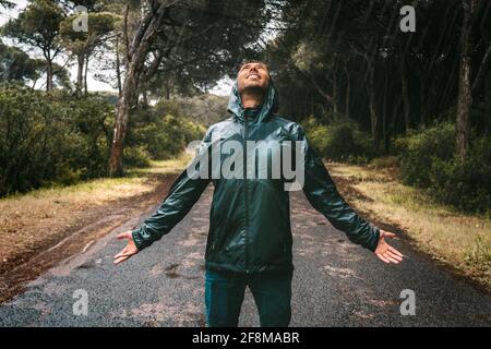 Junger Mann mit nassem Lächeln in einer durchnässten Jacke mit Kapuze, der den Regen genießt. Mann im Regenmantel stand bei starkem Regen. Schlechter Herbst, egal. Stockfoto