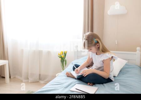 Ein kleines Mädchen sitzt auf dem Bett im Schlafzimmer und schreibt in einem blauen Buch. Bildungskonzept. Zu Hause Schulbildung. Hausaufgaben. Hochwertige Fotos Stockfoto