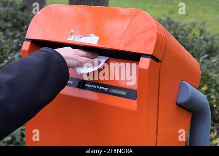Hand wirft Buchstaben in einen typisch holländischen orangefarbenen Briefkasten, der sich draußen in einer Straße entlang eines Parks befindet Stockfoto