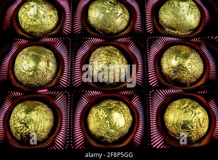 Eine Schachtel Pralinen in einer goldenen Folienverpackung. Süßigkeiten in glänzender Folie. Goldene Farbe. Süßes Dessert. Schachtel mit Süßigkeiten. Süßes Essen. Schwarzer Hintergrund. Packagin Stockfoto