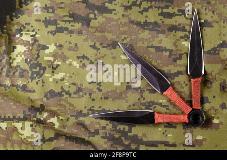 Schwarze Wurfmesser auf Camouflage-Tuch. Drei Spezialmesser mit roten Griffen gekreuzt. Militärisches Thema. Blick von oben in einem Winkel. Speicherplatz kopieren. Stockfoto