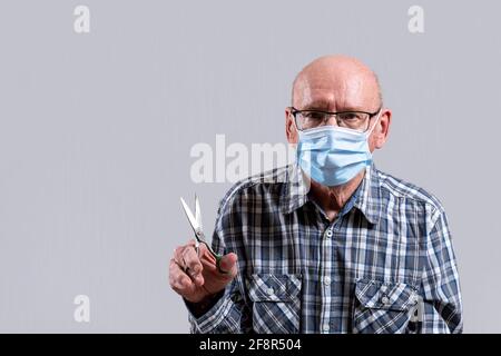 Alter kahler Mann mit Brille und medizinischer Maske mit Stahlschere in der Hand. Speicherplatz kopieren. Vorderansicht, horizontale Aufnahme im Innenbereich. Stockfoto