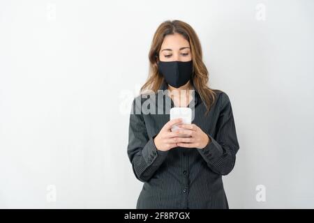 Studioaufnahme einer ernsthaften jungen Model-Frau im Business-Shirt Kleidung isoliert auf weißen Kopie Raum Hintergrund Blick auf Smartphone Das Tragen von medizinischen Prote Stockfoto