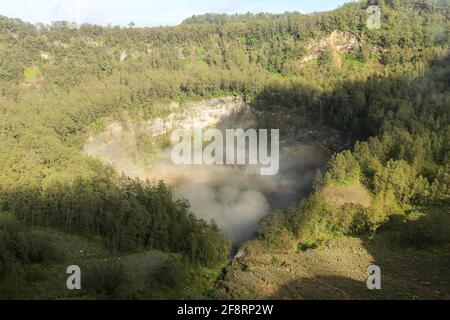 Das dunkelblaue Lagunenwasser eines der drei heiligen Seen von Kelimutu, Indonesien. Bäume umgeben den Krater, und neblige Wolken Rollen über den Krater Stockfoto