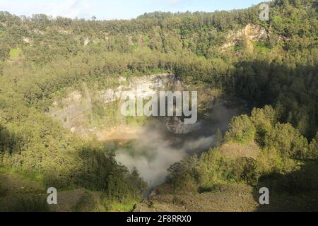 Das dunkelblaue Lagunenwasser eines der drei heiligen Seen von Kelimutu, Indonesien. Bäume umgeben den Krater, und neblige Wolken Rollen über den Krater Stockfoto