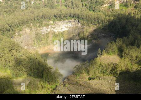 Das dunkelblaue Lagunenwasser eines der drei heiligen Seen von Kelimutu, Indonesien. Bäume umgeben den Krater, und neblige Wolken Rollen über den Krater Stockfoto