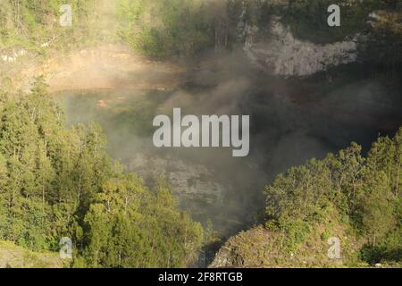 Das dunkelblaue Lagunenwasser eines der drei heiligen Seen von Kelimutu, Indonesien. Bäume umgeben den Krater, und neblige Wolken Rollen über den Krater Stockfoto