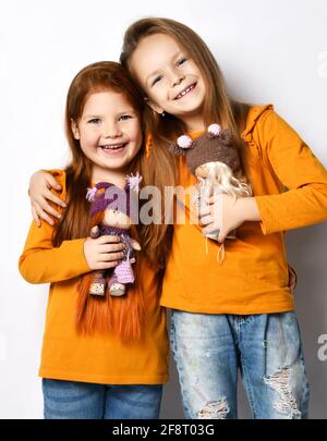 Glückliche kleine Mädchen Schwestern oder Freunde in Terrakotta gelb bequem langarm stehend umarmt und hält Spielzeugpuppen Stockfoto