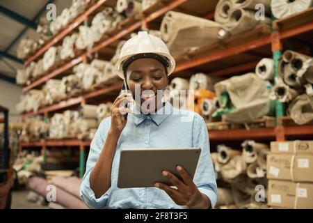 Lächelnde afrikanische Frau mit einem Tablet und Handy in einem Lager Stockfoto