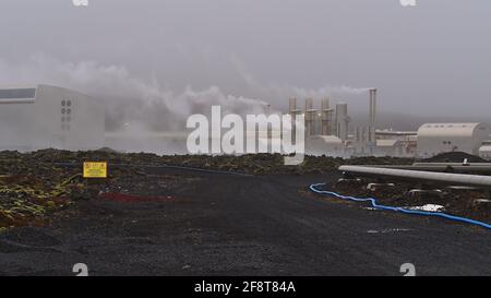 Blick auf das geothermische Kraftwerk Svartsengi mit dampfenden Kaminen und Warnschild neben moosbedecktem Lavafeld an bewölktem Tag in der Wintersaison. Stockfoto