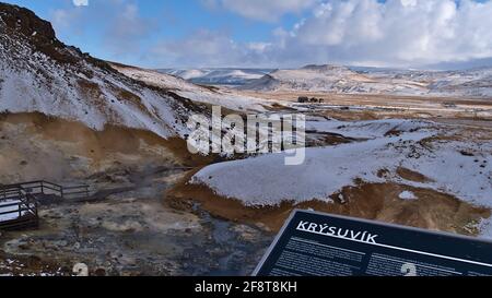 Blick über das geothermische Gebiet Seltún, Teil des vulkanischen Systems Krýsuvík, mit dampfenden heißen Quellen, Fumarolen und Informationstafel am bewölkten Wintertag. Stockfoto