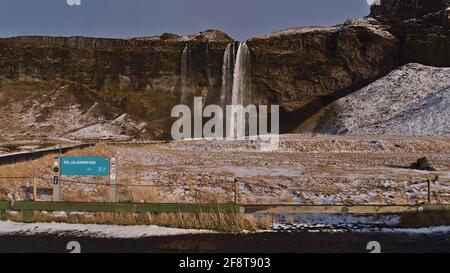 Schöne Vorderansicht des berühmten Wasserfalls Seljalandsfoss in der Wintersaison mit schneebedeckter Wiese und Namensschild. Stockfoto
