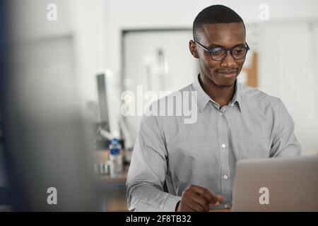 Lächelnder junger afrikanischer Geschäftsmann, der in einem Büro einen Laptop benutzt Stockfoto