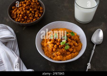 Dicke Kürbissuppe mit gebackenen Kichererbsen und Rettichsprossen auf einem Teller mit einem Glas Milch auf dem Tisch, leckeres gesundes Abendessen Stockfoto
