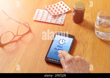 Pille Alarm App auf Handy, Handy oder Smartphone, mit Wasserglas, Brille, Medizin und die Hand einer älteren Frau. Stockfoto