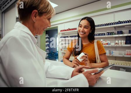 Glückliche leitende Apothekerin, die dem Bildschirm eines digitalen Tablets zeigt Junge Kunden halten eine Flasche Nahrungsergänzungsmittel in der Apotheke, während sie sich in der Apotheke befinden Ein Gespräch Stockfoto