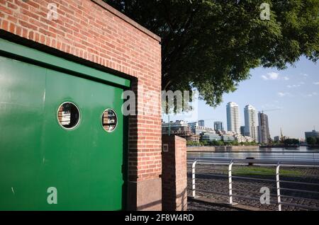 Kleines Gebäude aus roten Ziegeln mit grüner Metalltür in der Nähe des Flussufers. Üppige Vegetation und Hochhäuser im Puerto Madero-Viertel im Hintergrund. Stockfoto