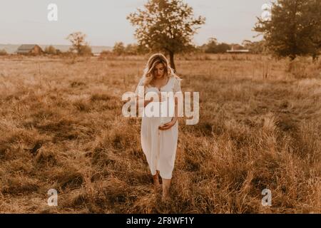 Kaukasische Schwangere junge blonde Frau in Baumwolle weißen Leinenkleid stehen zu Fuß Wiese auf trockenem Gras im Sommer bei Sonnenuntergang Natur. Mutter-zu-werden hält sie Stockfoto