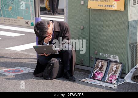 Japanischer Salaryman hockend auf der Straße mit seinem Laptop neben Kopien des Magazins „The Big Issue“ (verkauft von benachteiligten Menschen), Yoyogi, Tokio, Japan Stockfoto