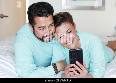 Glückliches und romantisches hispanische Paar, das sich ein Video auf der ansieht Telefon und Kuscheln im Bett Stockfoto