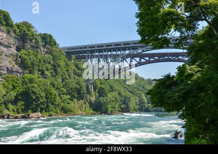 Sprudelnde Stromschnellen des Niagara River fließen über Steine und Felsen mit Brücke, Wald und Felswand im Hintergrund bei sonnigem Wetter Stockfoto