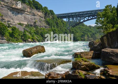 Sprudelnde Stromschnellen des Niagara River fließen über Steine und Felsen mit Brücke, Wald und Felswand im Hintergrund bei sonnigem Wetter Stockfoto