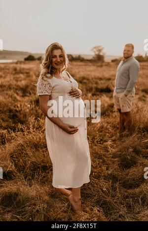 Glückliche Familie schwanger kaukasischen blonde Frau mit Maulwürfe in weißem Baumwollkleid Spaziergänge mit Mann auf der Wiese im Sommer. Mann Licht natürliche Kleidung und sch Stockfoto