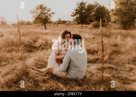 Die Schwangeren sitzen in den Armen ihres Mannes und küssen und umarmen ihn. Glückliche Familie Ruhe Natur umarmt Küsse im Sommer bei Sonnenuntergang. Zukünftige Mutter kaukasischen w Stockfoto