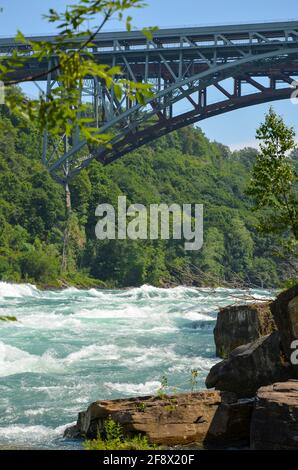 Sprudelnde Stromschnellen des Niagara River fließen über Steine und Felsen mit Brücke, Wald und Felswand im Hintergrund bei sonnigem Wetter Stockfoto