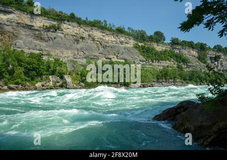 Sprudelnde Stromschnellen des Niagara River, die über Steine und Felsen fließen Mit Wald- und Felswand im Hintergrund bei Sonnenschein Wetter Stockfoto