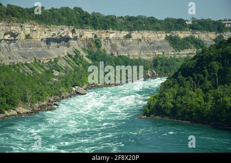 Sprudelnde Stromschnellen des Niagara River, die über Steine und Felsen fließen Mit Wald- und Felswand im Hintergrund bei Sonnenschein Wetter Stockfoto