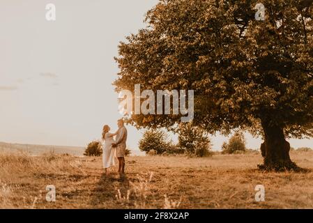 Ehemann umarmt schwanger Frau mit großen Bauch. Glückliche Familie in der Natur ruht umarmt Küsse im Sommer bei Sonnenuntergang. Paar auf dem Hintergrund von großen einsamen Baum. Stockfoto