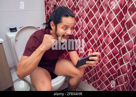 Ein junger lateinischer Mann lächelte auf der Toilette und benutzte ein Smartphone im Badezimmer. Stockfoto