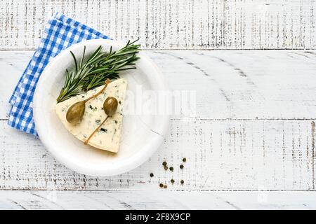 Stücke Blauschimmelkäse oder Brie-Käse in einer Schüssel mit Rosmarin, Kapern und Pfeffer. Auf weißem Holz alten Hintergrund. Draufsicht. Freier Speicherplatz für Ihren Text. Stockfoto