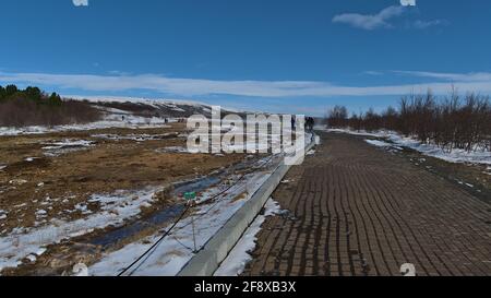 Touristen, die am sonnigen Wintertag auf einem Fußweg in Geysir, einem geothermischen Gebiet, am Goldenen Kreis, mit Warnschild und dampfenden heißen Quellen wandern. Stockfoto