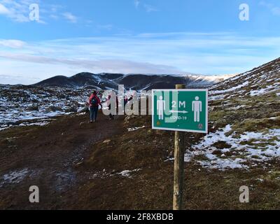 Menschen wandern auf dem Weg zum kürzlich erupten Vulkan am Fagradalsfjall mit Warnschild, das daran erinnert, wegen Covid-19 zwei Meter Abstand zu halten. Stockfoto