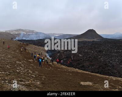 Blick auf den Vulkanausbruch am Fagradalsfjall in den Geldingadalir-Tälern mit Menschen, die an einem schwarzen, glühenden Lavafeld mit Rauch vorbeiziehen. Stockfoto