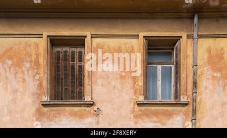 Verlassene Fenster mit offenen und geschlossenen Fensterläden an der Fassade eines verlassenen Wohngebäudes. Zwei Fenster auf schmutzigen braunen Wandhintergrund. Stockfoto